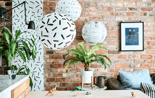 Three round black and white patterned lamps hang over a countertop with toy figures, in a kitchen/diner with a brick wall.