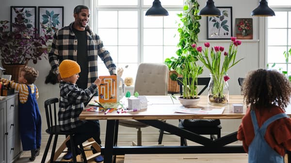 Three small children and their father go happily about their business in a dining area with a SKOGSTA table.