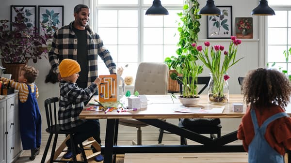 Three small children and their father go happily about their business in a dining area with a SKOGSTA table.