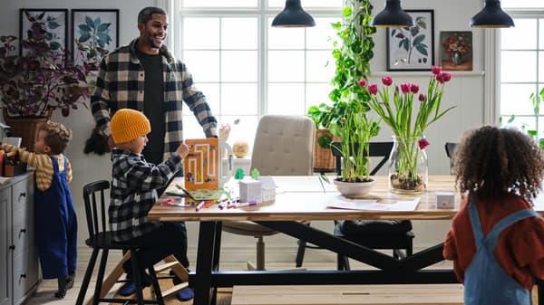 Three small children and their father go happily about their business in a dining area with a SKOGSTA table.