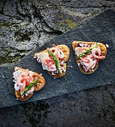 Three small waffle-like appetizers topped with shrimp, asparagus, cheese shavings, and sauce, arranged on a dark stone slab with a textured background.