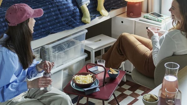Three students in a dorm room gathered around an IKEA PS 2026 side table, with a KULGLASS speaker lamp in the background.