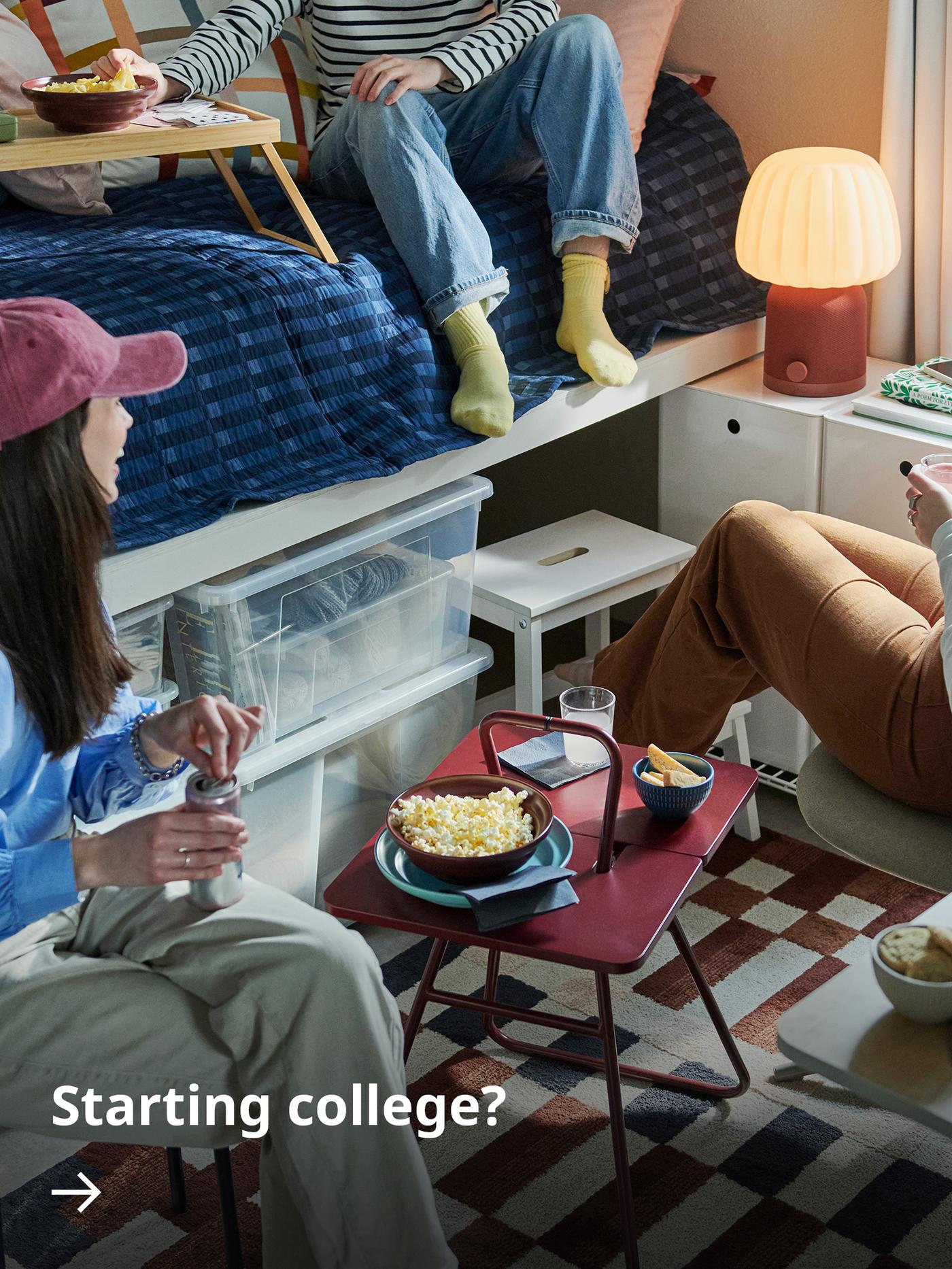 Three students in a dorm room gathered around an IKEA PS 2026 side table, with a KULGLASS speaker lamp in the background.