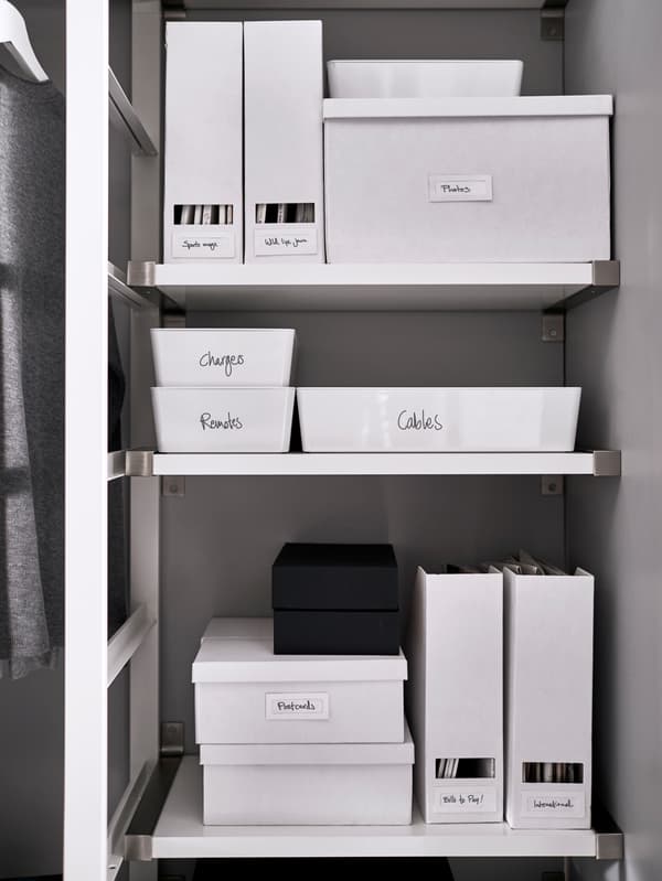 Three white shelves that hold white boxes and magazine holders with handwritten labels.