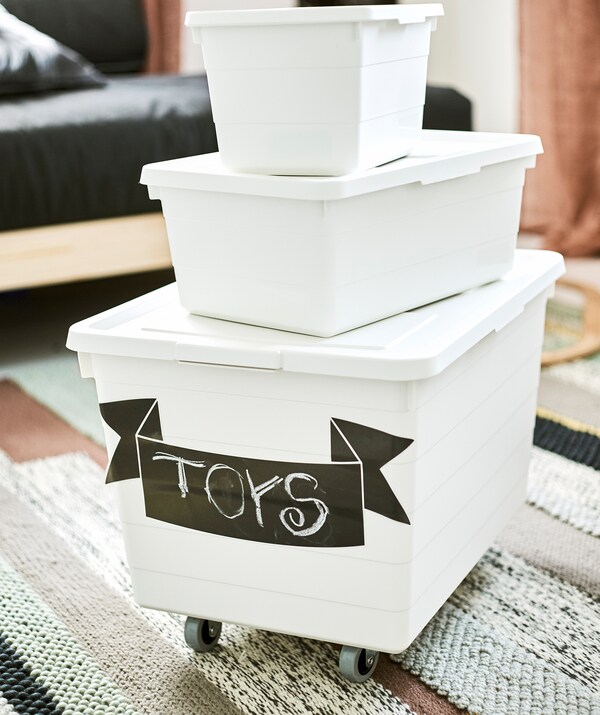 Three white storage boxes of different sizes piled up on a striped rug. The bottom box is labelled toys and has wheels.