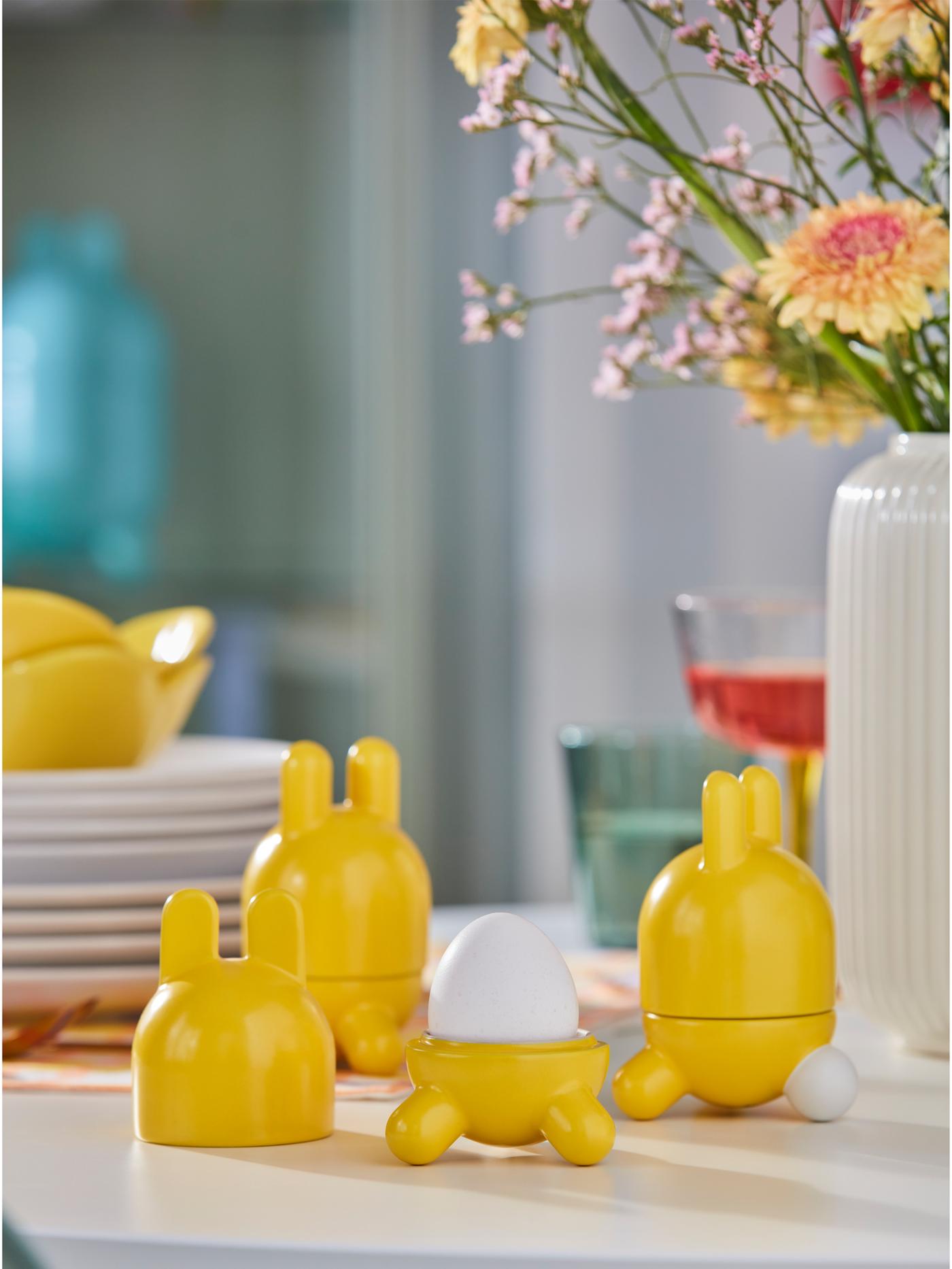 Three yellow TJÄRLEK egg cups are placed on a white tabletop. The cup in the middle has no lid on, and there is an egg in it.
