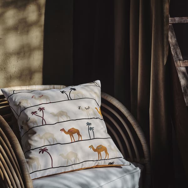TILLTALANDE cushion cover with hand-embroidered camels and palm trees against a white background placed in a rattan chair.