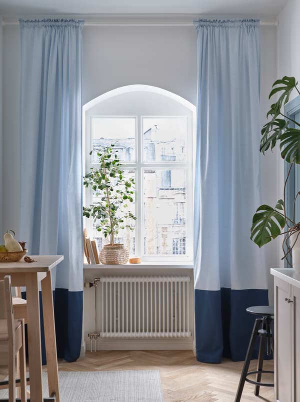 Tingkap dapur yang bulat dengan pokok besar di ambang tingkap dan langsir penghalang cahaya BENGTA berwarna biru lembut di kedua-dua belah.