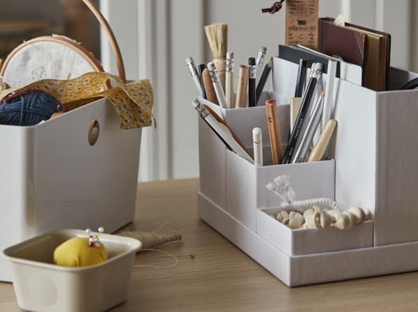 TJENA desk organiser on a table top with stationery items and craft supplies organised in each compartment. A KUGGIS and NOJIG storage box sit next to the organiser with other craft materials. 