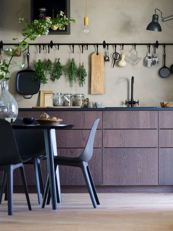 Tmavohnedá kuchyňa s čiernou pracovnou doskou a čiernym drezom. Nad ňou je čierny závesný úložný systém HULTARP na béžovej stene. V popredí čierny stôl a čiernomodré stoličky BALTSAR.