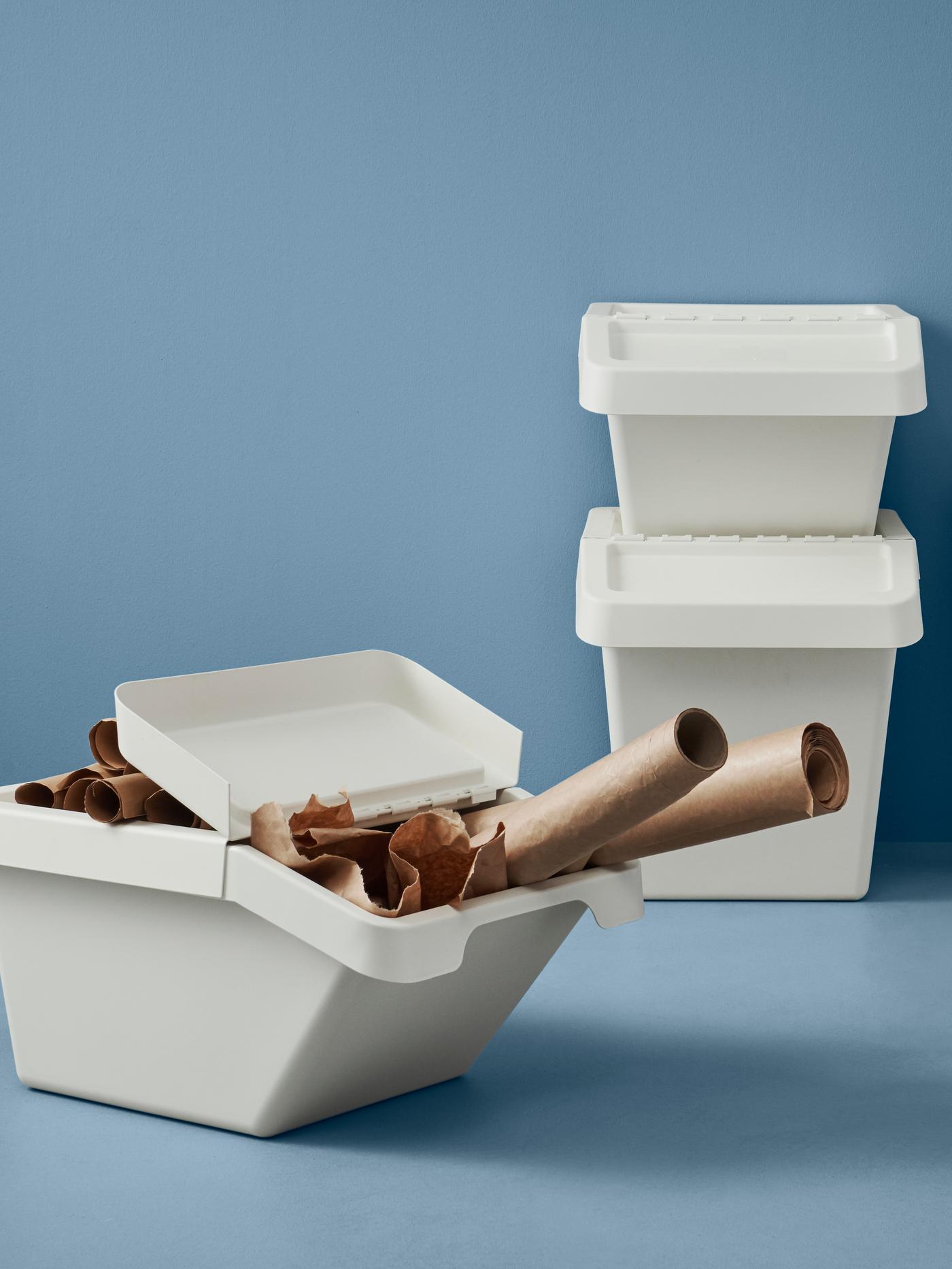 To affaldssorteringsspande står ovenpå hinanden en tredje har et halvt åbent låg hvor der stikker ruller af brun pap papir ud af de hele står i et tomt rum med en blå baggrund.