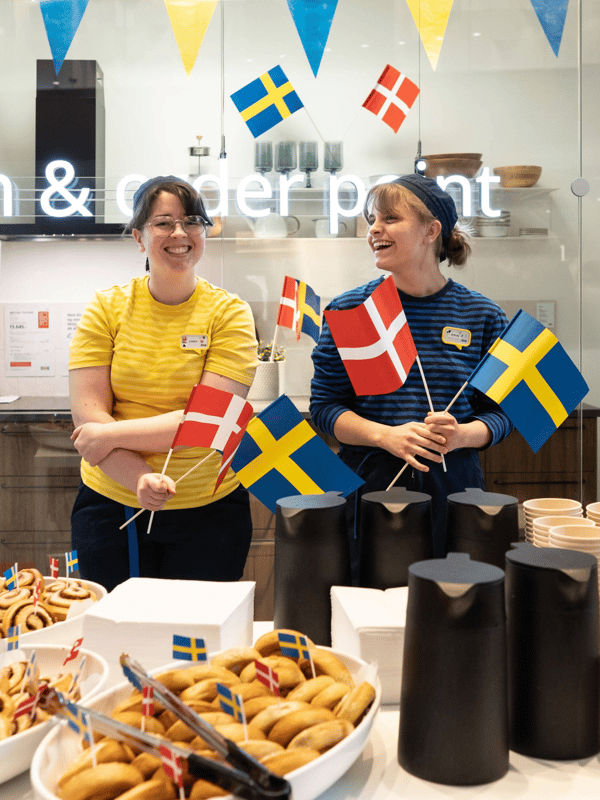 To IKEA medarbejdere i deres uniformer står og flager med det svenske flag foran et bord dækket med kanelsnegle og kaffekander.
