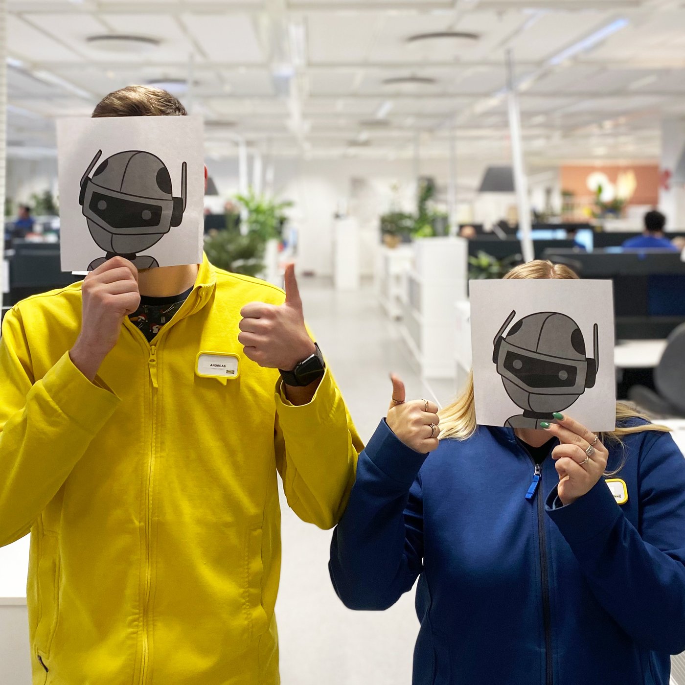 To IKEA medarbejdere i uniformer holder billeder af en robot op foran deres ansigt mens de viser tommel up med den anden hånd.
