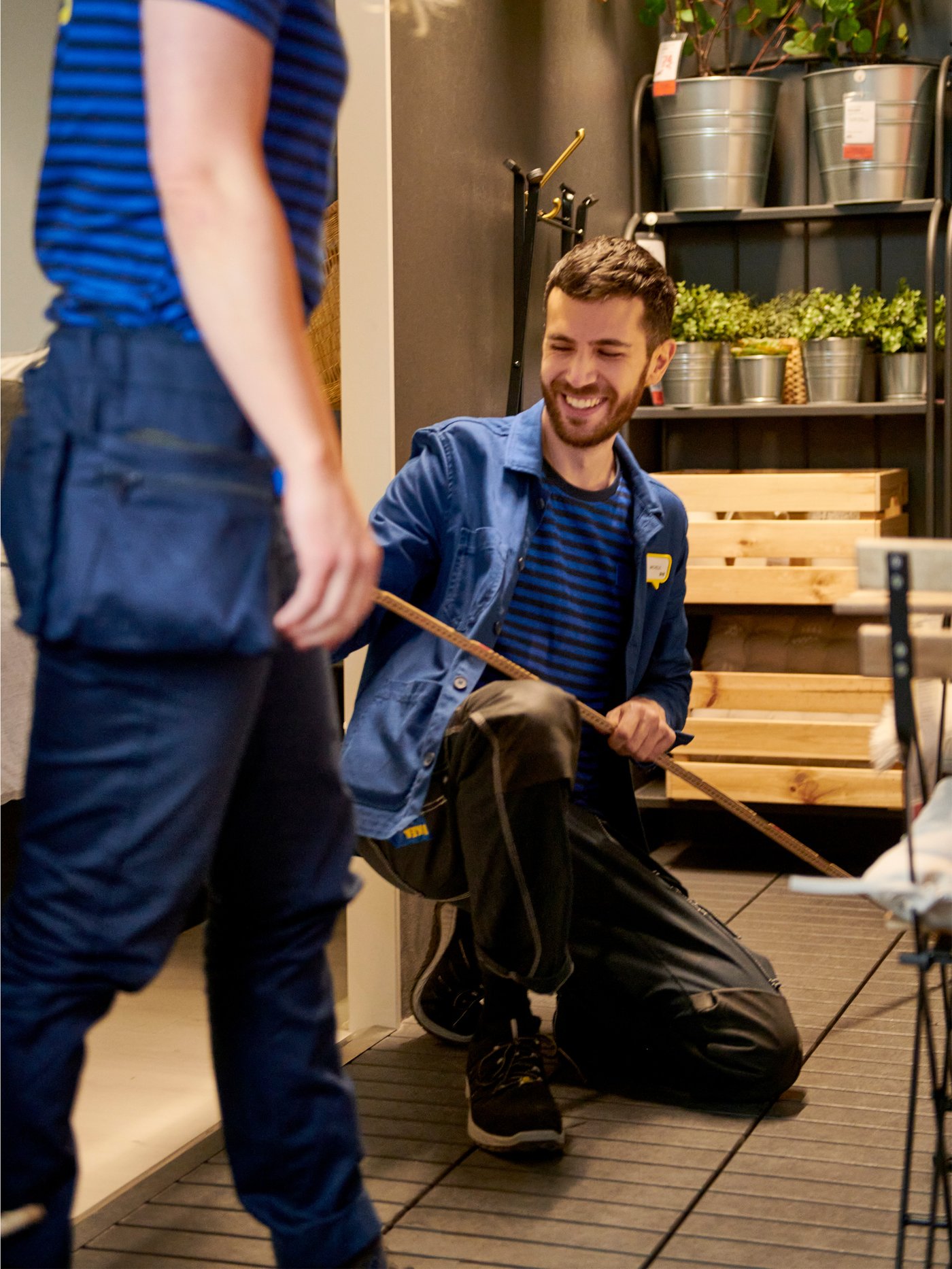 To IKEA medarbejdere står i en udstilling den ene sidder på knæ med en tommestok og siler mens den anden kigger væk fra kameraet.