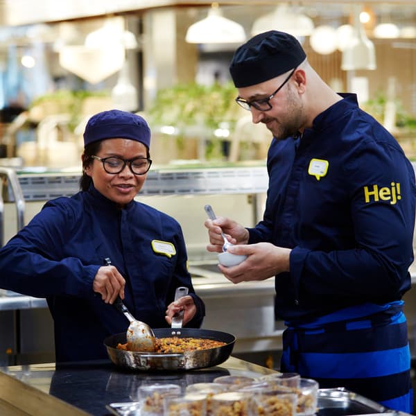 To IKEA medarbejdere står og tilbereder mad på en kogeplade den ene med briller rør i en pande mens den anden holder en lille skål og en ske i hånden.