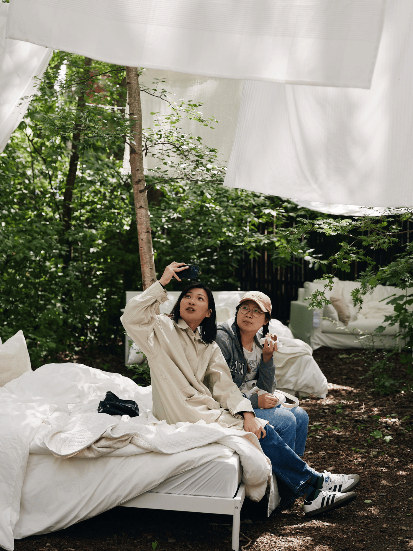 To kvinder sidder på en seng der står udenfor den ene kvinder er ved at tage et selvportræt med sin telefon og sengene er redt med hvidt sengetøj dyner og puder.