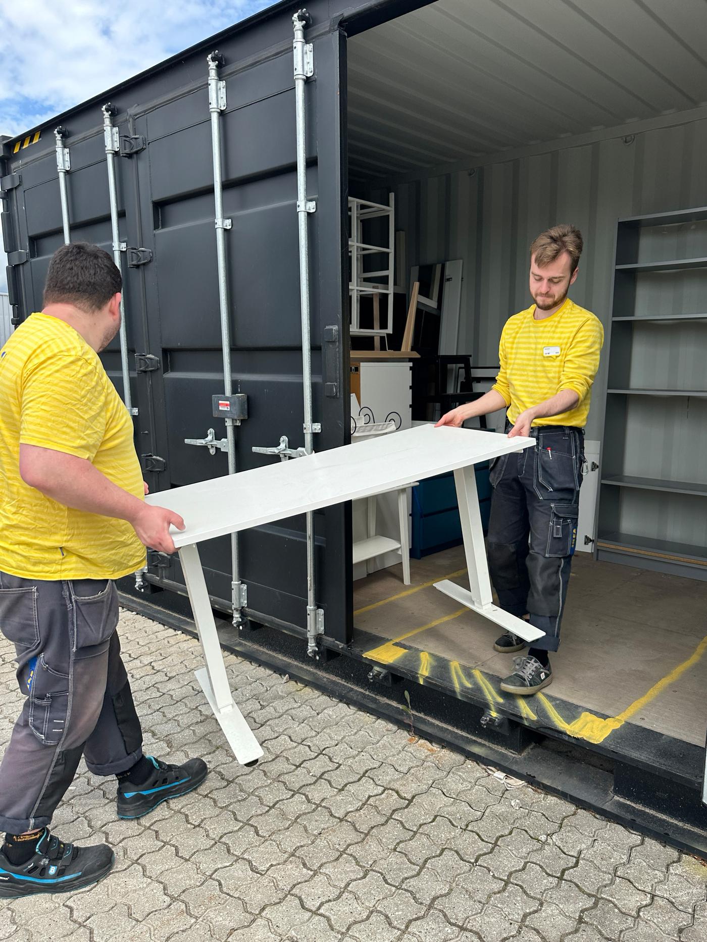 To mænd i IKEA uniformer bærer et hvidt skridbord ind i en stor container, hvor der også står andre møbler.