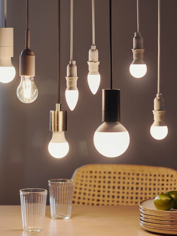 Többféle méretű és formájú SOLHETTA LED izzót látunk egy asztal fölött. Az asztalon két VARDAGEN pohár áll néhány egymásra rakott bézs GLADELIG tál társaságában.