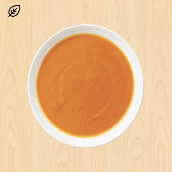 Top-down view of a white bowl filled with smooth orange soup on a light wood table; a leaf icon suggests a natural or vegan dish.