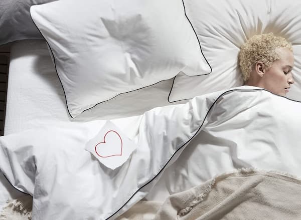 Top down view of a woman asleep in bed with blankets over her and a paper with a heart drawn on it next to her.