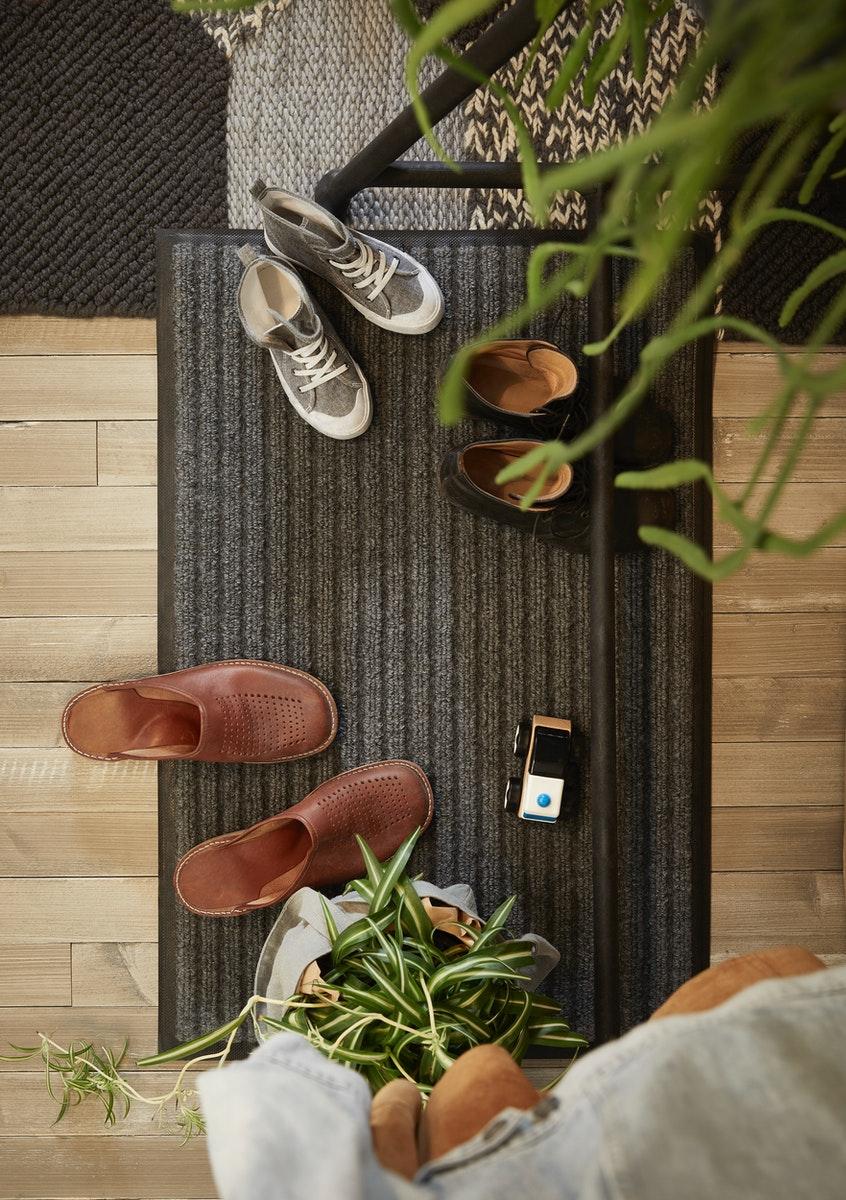 Top-down view of shoes on a ribbed dark brown doormat near a light wooden floor and a plant.