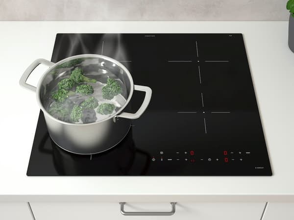 Topf mit kochendem Brokkoli auf einem schwarzen Induktionskochfeld mit Touch-Bedienung und Markierungen für die Heizzonen; Dampf steigt aus dem Topf auf. Das Kochfeld ist in eine weiße Arbeitsplatte mit darunterliegender Schublade eingebaut.