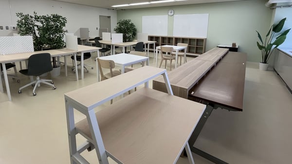 観葉植物と仕切り付きデスクが並ぶ明るい教室。木製テーブルと収納で、集中しやすい自習・ワークスペースを演出。