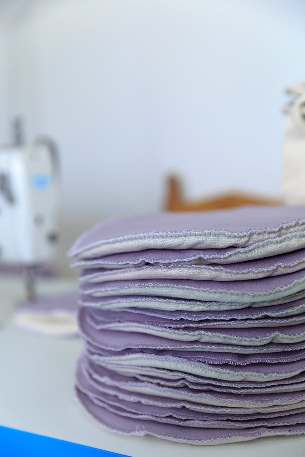 Tortilleros lilas apilados, sobre la mesa y al fondo una máquina para coser.