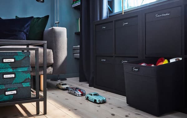 Toy cars lined up on light wood flooring in a living room with black furniture and storage boxes for toy organisation.