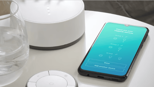 Trådfri gateway on a table next to a mobile phone with the IKEA Home Smart 1 app open showing on the screen