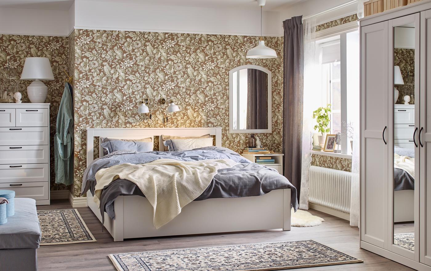 Traditional beige, blue and white bedroom with floral wall paper and white SONGESAND bed, bedside table and chest-of-drawers.