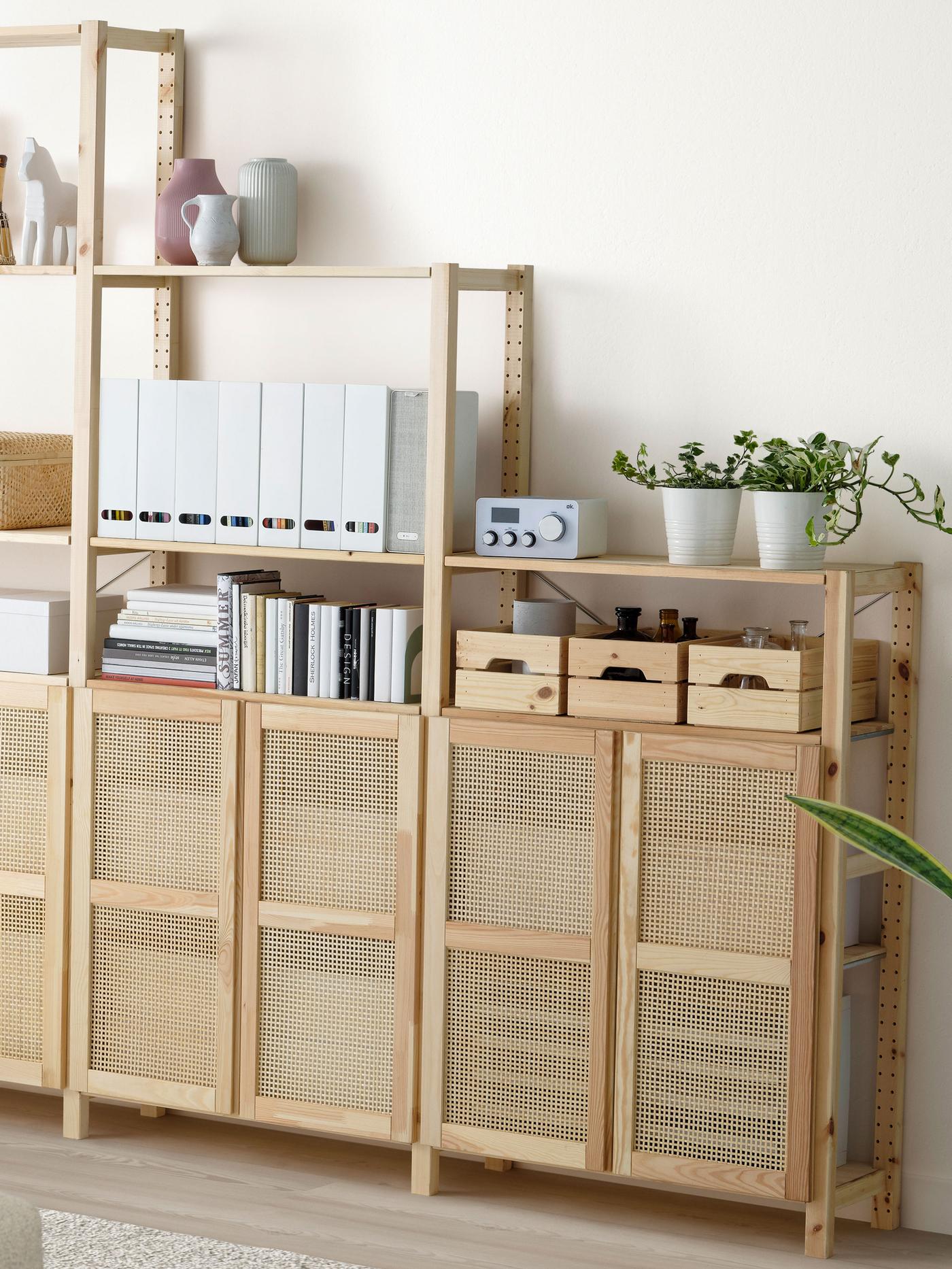 Trähylla med flera fack och hyllplan, fylld med böcker, pärmar, små förvaringslådor, krukväxter och dekorativa föremål. De nedre facken är delvis täckta med dörrar i flätad rotting.