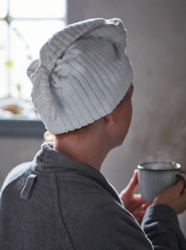 ผู้หญิงสวมหมวกผ้าคลุมผม TRÄTTEN/เทรทเตน และสวมเสื้อคลุมอาบน้ำ ถือแก้วชาอยู่ในห้องน้ำ