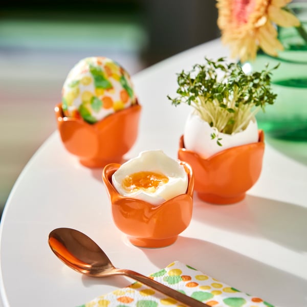 Tre äggkoppar i orange form av blommor. Ett ägg är dekorerat, ett annat är kokt och halvt öppet, och det tredje innehåller krasse. En sked och en färgglad servett ligger bredvid.