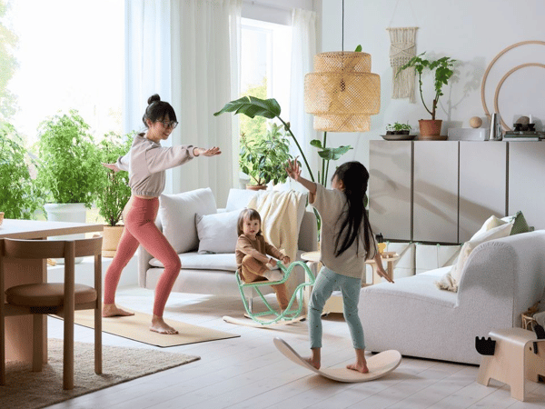 Tre barn leker i ett ljust vardagsrum, en gör gymnastik, en gungar på en gungälg och en på en bräda i ljust trä.