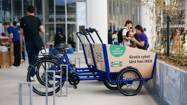 Tre blå IKEA elladcykler der er gratis at låne står ude foran IKEA København varehuset og i baggrunden sidder flere mennesker og slapper af.