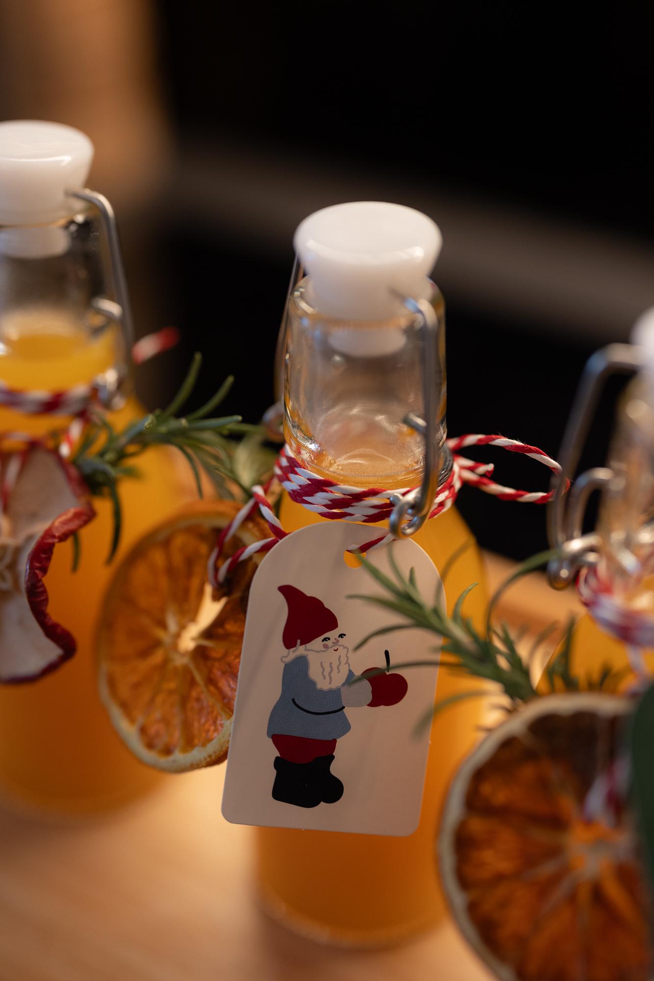 Tre bottiglie di vetro riempite con una bevanda dorata, decorate con fette d'arancia, rosmarino e festosi cartellini con uno gnomo.