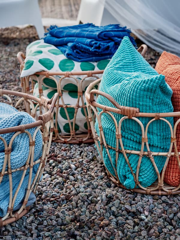Tre cesti SNIDAD con cuscini colorati e biancheria posti su un terreno di sassi.