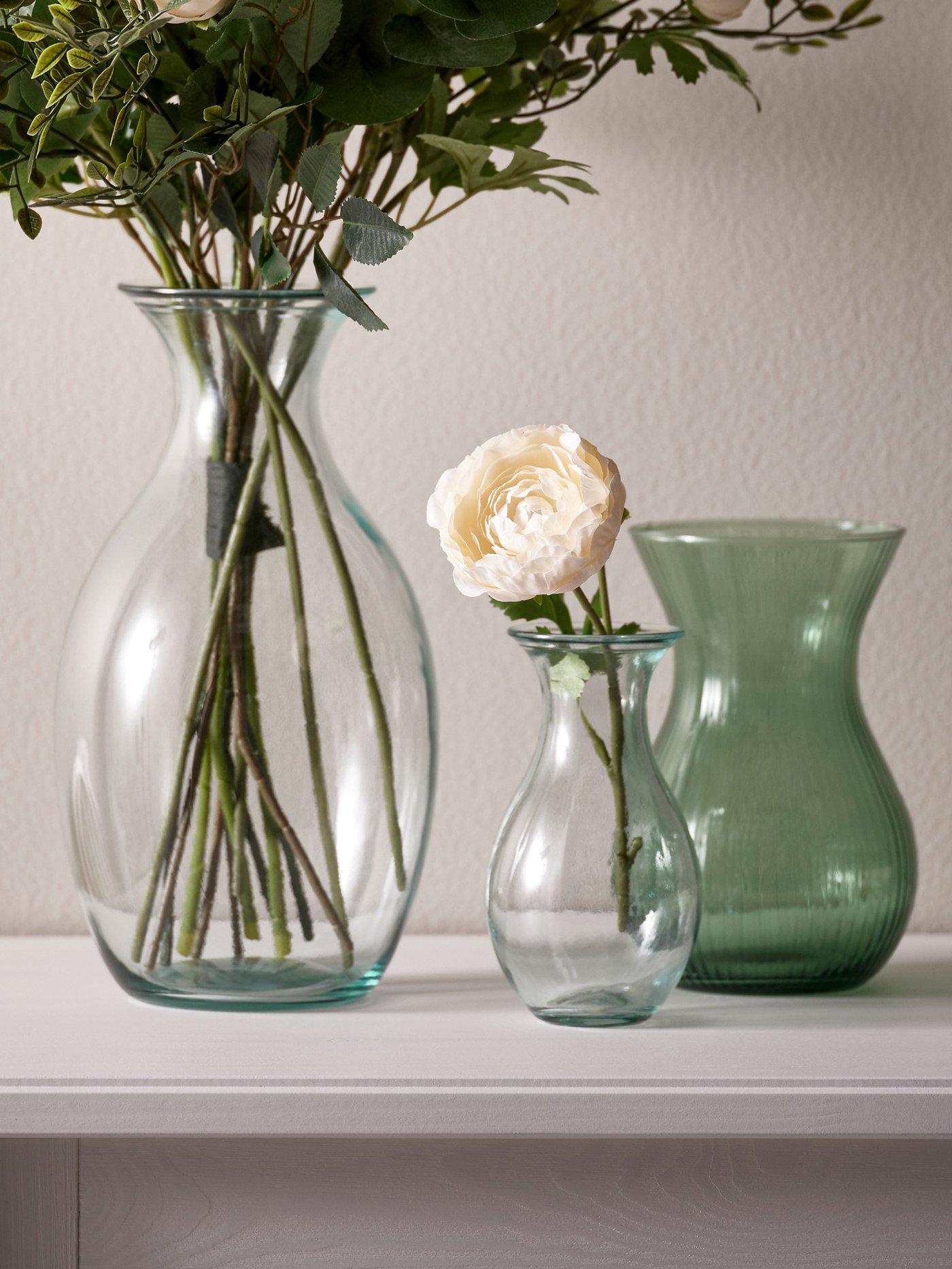 Tre genomskinliga glasvaser i olika former med gröna nyanser, fyllda med blommor, placerade på en vit hylla.