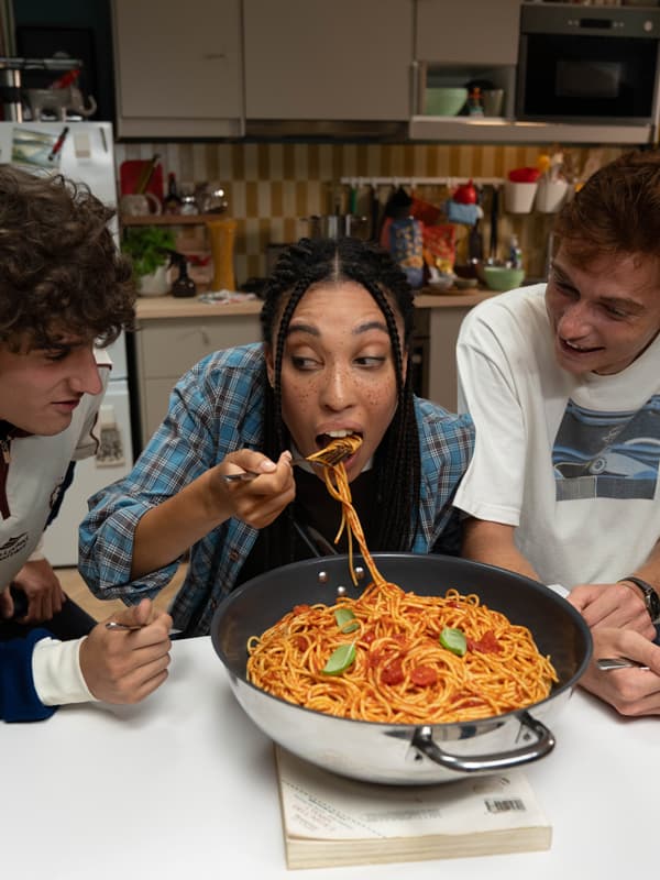 Tre giovani amici mangiano con allegria un piatto di spaghetti direttamente dalla pentola, condivisa al centro del tavolo.