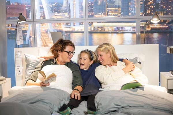 Tre glada personer som halvligger i en säng varav den ena löser en bok för de andra. 