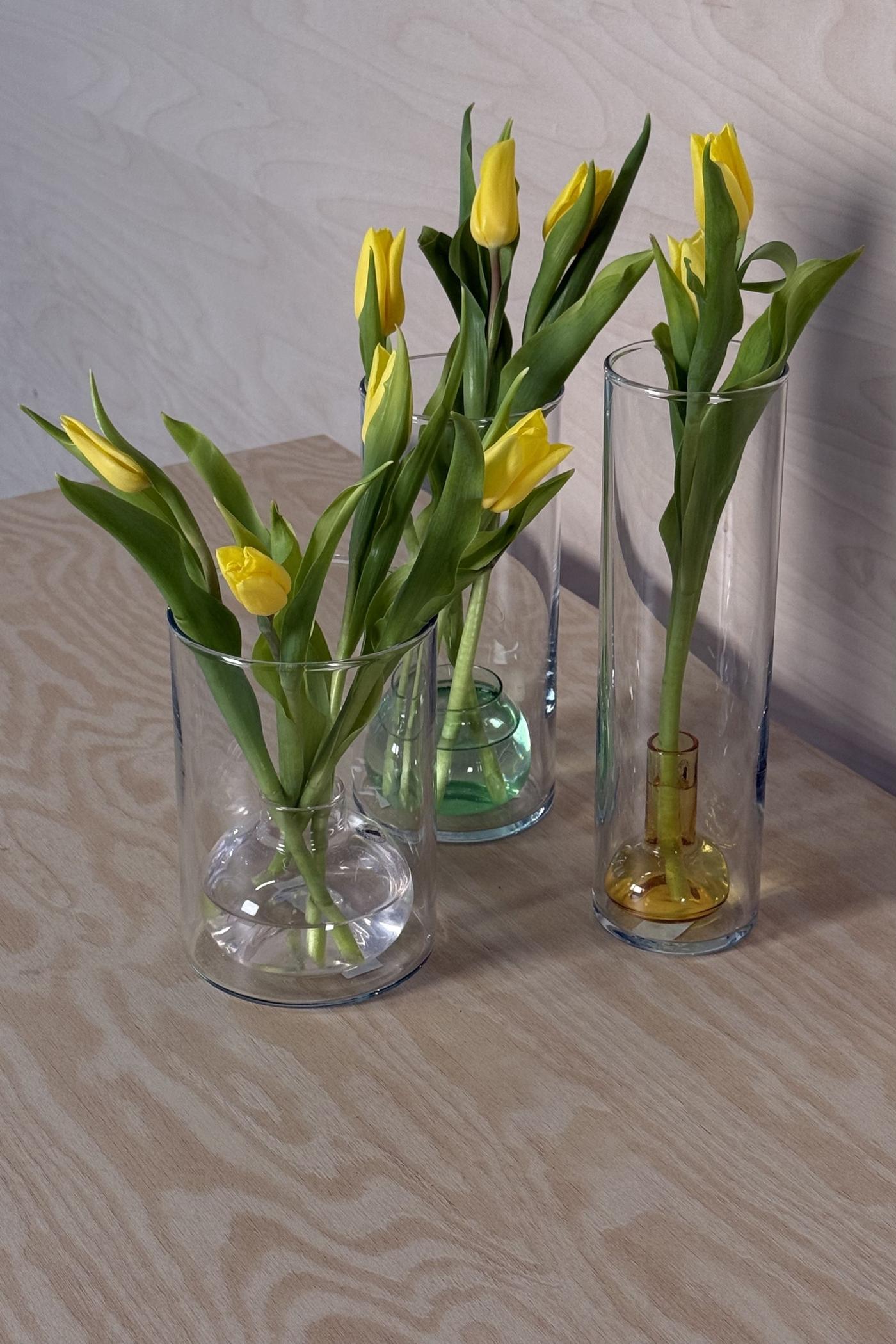 Tre höga glasvaser med mindre vaser inuti står på ett ljust träbord, fyllda med gula tulpaner.