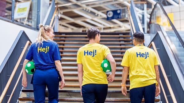 Tre IKEA medarbejdere med ryggen til, en i blå uniform og to i gule uniformer, går op af en bred trætrappe i et IKEA varehus.