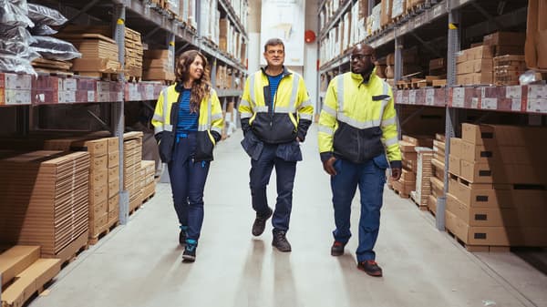Tre lagerarbejdere i synlige jakker går ned ad en gang i IKEAs distributionscenter. Hylder fyldt med kasser og pakkede varer langs væggene. De ser ud til at snakke og fokusere på deres opgaver som ledertrainee.