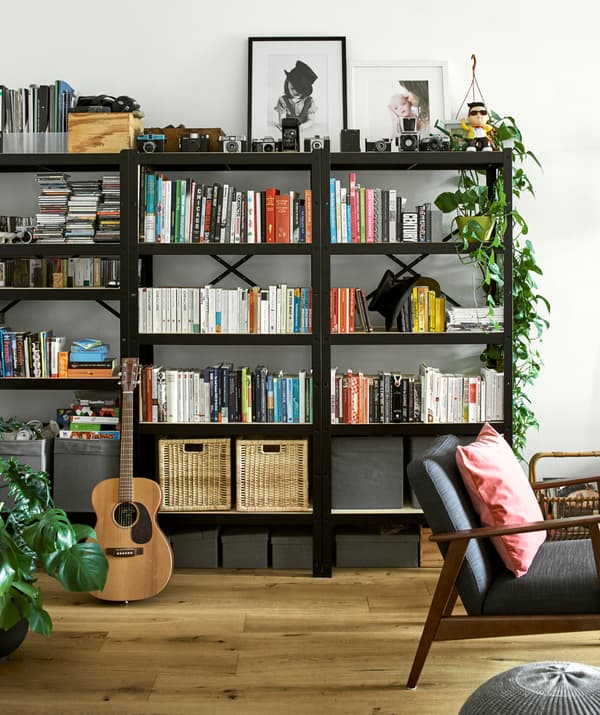 Tre librerie nere con libri disposti per colore, cestini in rattan, foto incorniciate, fotocamere vintage e piante.
