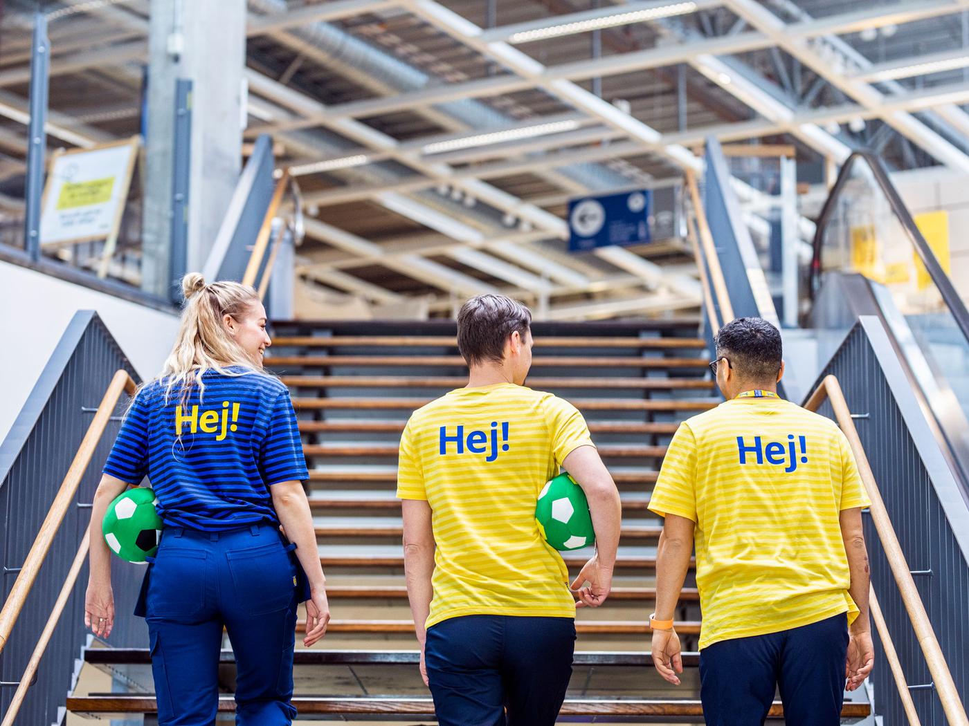 Tre personer går op ad en trappe inde i en moderne IKEA varehus. De er iført knaldblå og gule skjorter med "Hej!" trykt på bagsiden. Hver person holder en grøn fodbold, der repræsenterer IKEAs værdier og skaber et muntert, venligt miljø.