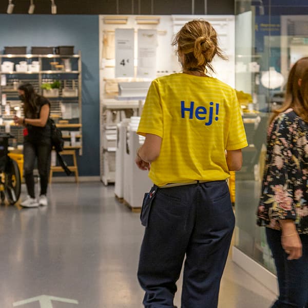 Treballadora d’IKEA amb una samarreta groga on hi diu “Hej!”, caminant per la botiga. Altres persones compren al seu voltant.