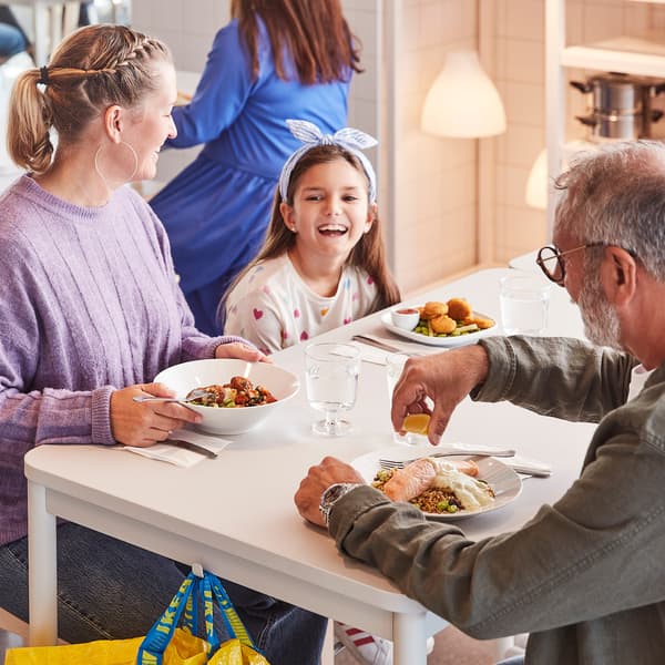 Trei persoane stau la o masă albă într-un restaurant, mâncând. O femeie în pulover mov și o fetiță cu bandană albastră sunt pe partea stângă, iar un bărbat cu barbă și ochelari este pe partea dreaptă. În fundal, o altă femeie în rochie albastră.