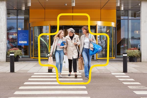 Trei persoane traversează o trecere de pietoni în fața unui magazin cu pereți galbeni și intrare rotativă, purtând pungi de cumpărături.