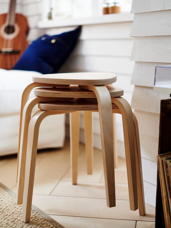 Trei scaune KYRRE stivuite lângă un perete cu lambriu alb, la câțiva metri de o canapea albă, cu o chitară pe ea.